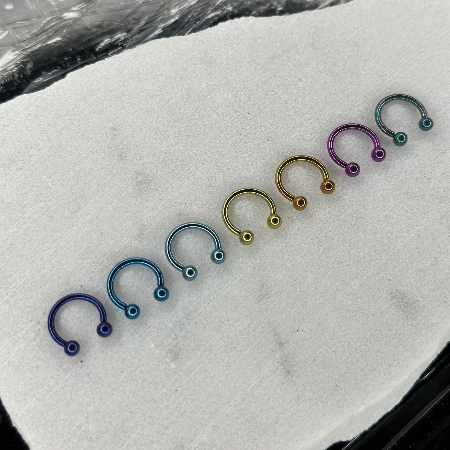 Una fila di gioielli da piercing in titanio anodizzato a forma di ferro di cavallo (circular barbell). I piercing mostrano una gamma di colori personalizzati, inclusi blu, oro, viola e verde, disposti su una salvietta sterile