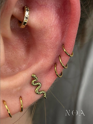 Primo piano di un 'curated ear' con gioielli piercing in oro. In evidenza un piercing a forma di serpente (snake) sulla cartilagine, multipli anelli dorati sull'helix e un anello con zirconi nel conch. Stile di NOA INK.