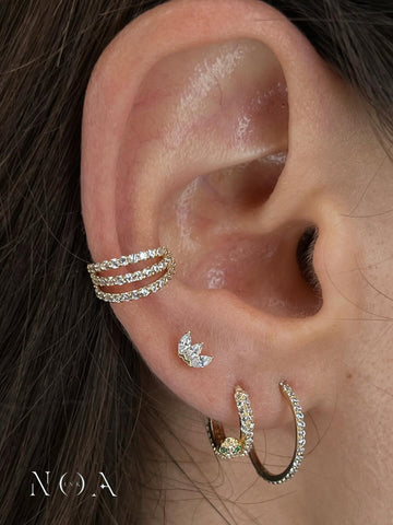 Primo piano di un 'curated ear' con gioielli in oro e zirconi. In evidenza un ear cuff a triplo pavé sulla cartilagine, un piercing 'flat' a corona (marquise cluster) e due orecchini a cerchio pavé nel lobo. Stile elegante di NOA INK