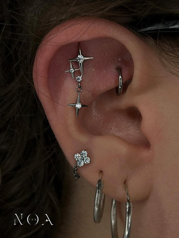 Primo piano di un 'curated ear' con un piercing industriale/helix lungo a stella cadente (dangle star) e un piercing al lobo a forma di fiore a tre petali con zirconi. Gioielli in titanio o PVD nero/gunmetal, stile galassia/celestiale di NOA INK.