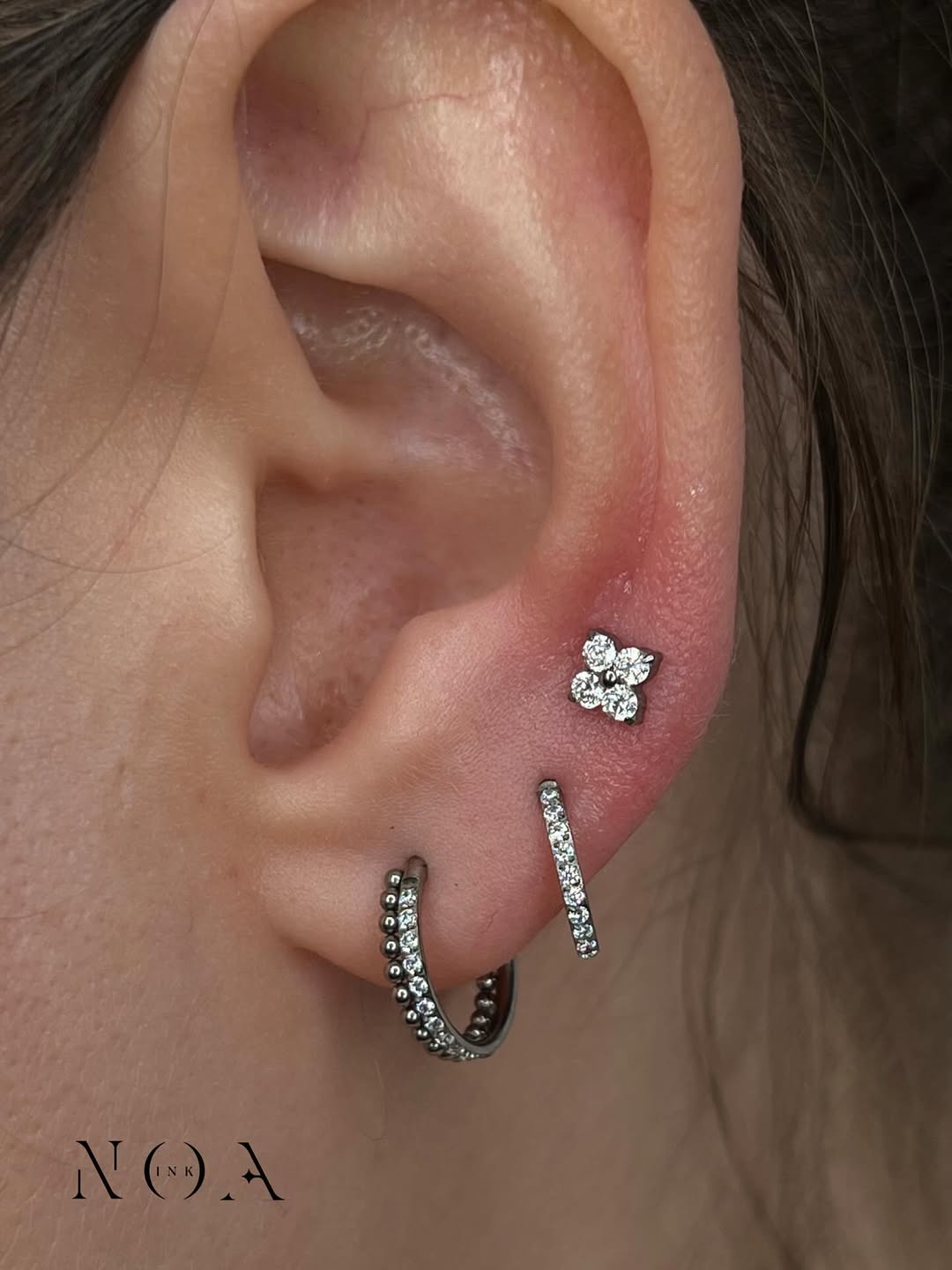 Primo piano di un orecchio con un piercing helix 'flat' a forma di fiorellino con zirconi bianchi e due orecchini a cerchio pavé nel lobo. I gioielli sono in titanio chirurgico o oro bianco/argento, stile curato da NOA INK.