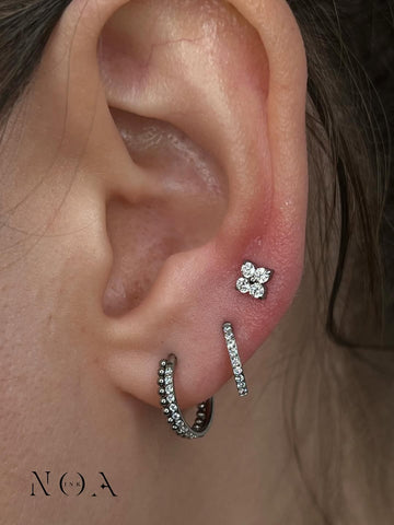 Primo piano di un orecchio con un piercing helix 'flat' a forma di fiorellino con zirconi bianchi e due orecchini a cerchio pavé nel lobo. I gioielli sono in titanio chirurgico o oro bianco/argento, stile curato da NOA INK.