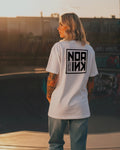 Donna con tatuaggi indossa la Icon Drop T-Shirt bianca di Noa Ink in un contesto urbano al tramonto. La maglietta oversize presenta il logo Noa Ink sul retro. Outfit casual con jeans strappati e sneakers bianche, illuminato da luce calda dorata.