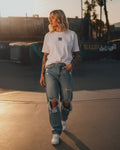 Donna con tatuaggi indossa la Icon Drop T-Shirt bianca di Noa Ink in un contesto urbano al tramonto. La maglietta oversize presenta il logo minimalista Noa Ink sul petto. Outfit casual con jeans strappati e sneakers bianche, illuminato da luce calda dorata.