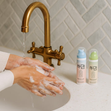 Due flaconi Hayan (Soluzione Salina e Schiuma Detergente) per cura piercing, affiancati a un lavandino di marmo con rubinetto dorato, mentre una persona si lava le mani