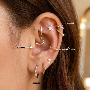 Guida visiva alle misure dei piercing all'orecchio: esempio di curated ear con gioielli in oro e brillanti su helix, conch, tragus e lobo, con annotazioni delle taglie in mm (6mm, 8mm, 10mm) per la scelta del gioiello perfetto