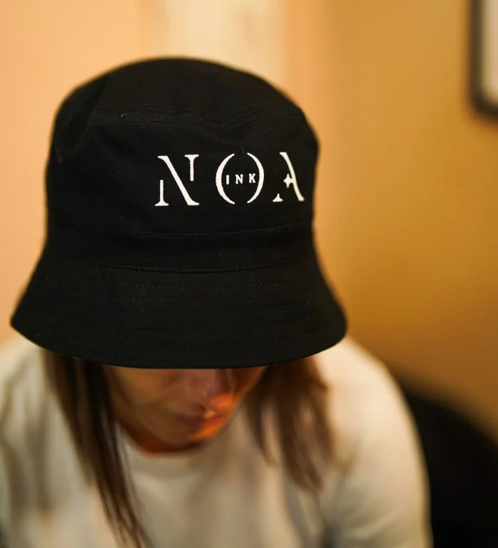 Cappellino nero Noa Ink con logo bianco ricamato, indossato da ragazza e ragazzo in look streetwear. Design unisex in cotone, con visiera curva e stile minimal urbano.