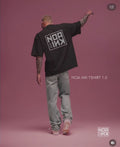 Uomo con tatuaggi indossa la Noa Ink icon drop T-Shirt nera oversize, con maxi logo quadrato bianco sul retro. Sfondo rosa minimal, look streetwear con jeans chiari e sneakers. Maglietta unisex firmata Noa Ink, dallo stile urbano e contemporaneo.